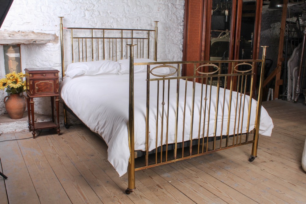 An antique brass king-size bed frame featuring vertical spindles, decorative oval motifs, and four tapered posts on castors.