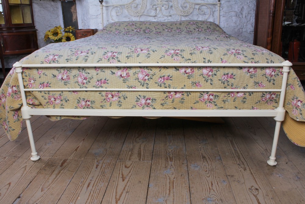 A king-size cream painted forged iron bedstead featuring an arched scrollwork headboard with a central decorative crest and a low footboard. View II