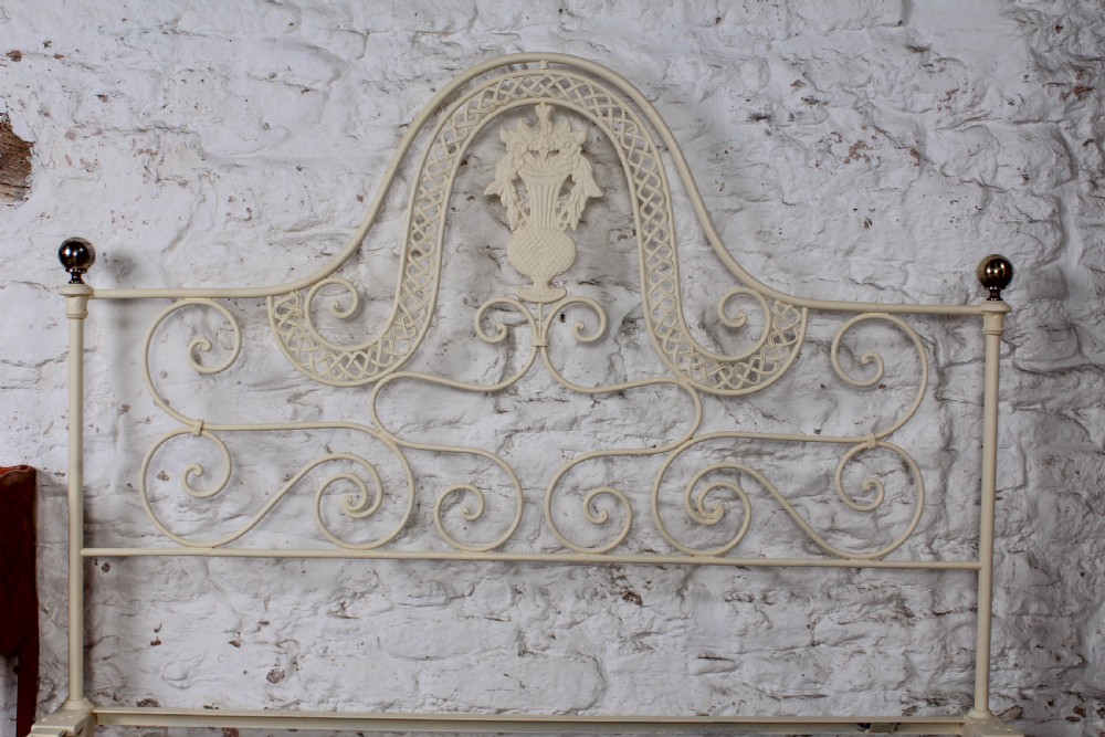 A king-size cream painted forged iron bedstead featuring an arched scrollwork headboard with a central decorative crest and a low footboard. View V