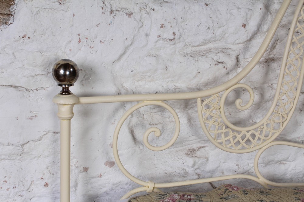 A king-size cream painted forged iron bedstead featuring an arched scrollwork headboard with a central decorative crest and a low footboard. View VI