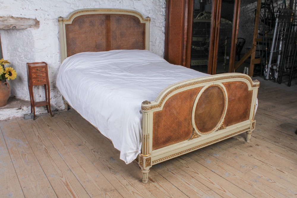 A French Louis XVI style king size bedstead featuring cream painted and gilded carved wood with natural woven cane panels on the headboard and footboard.