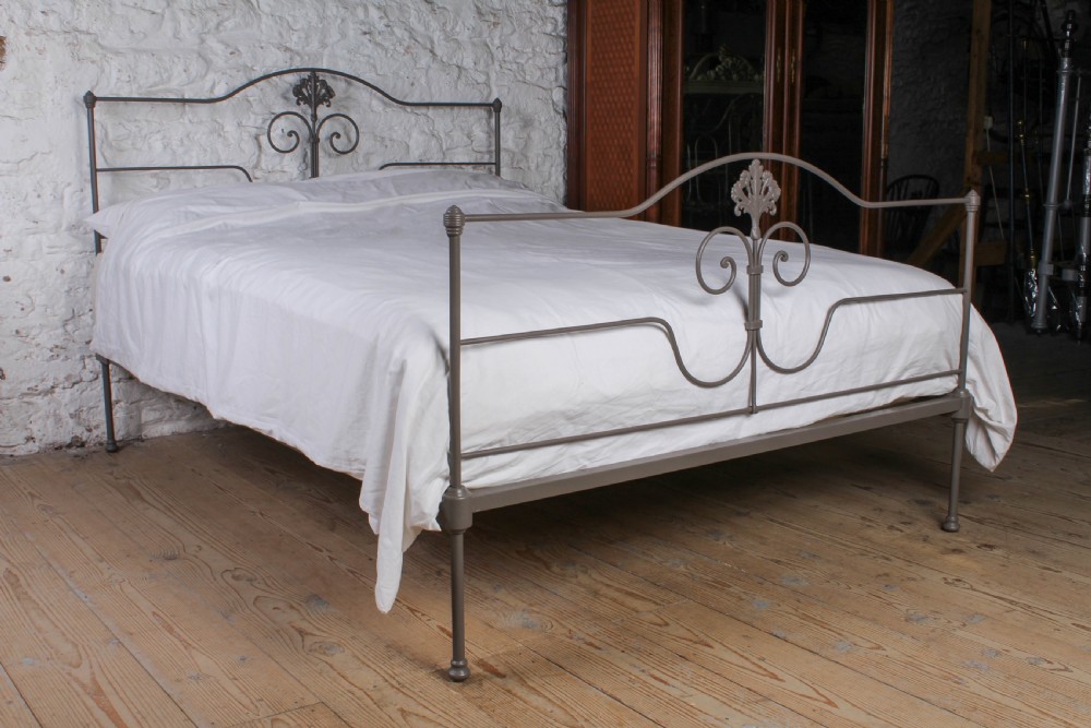 A king size Portuguese iron bed frame painted in a neutral taupe colour, featuring decorative scrollwork and a central shell motif on both the headboard and footboard.