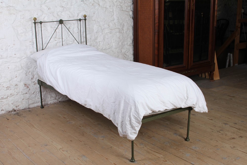 A small Victorian single platform bed constructed from dark green painted iron with brass corner finials.