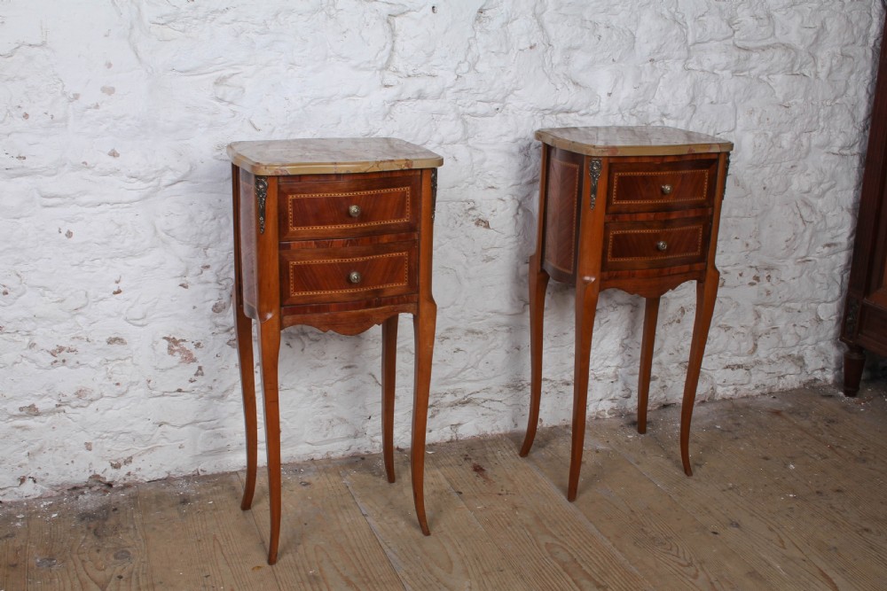 A pair of slender French wooden bedside tables featuring variegated marble tops, two inlaid drawers, and high cabriole legs.
