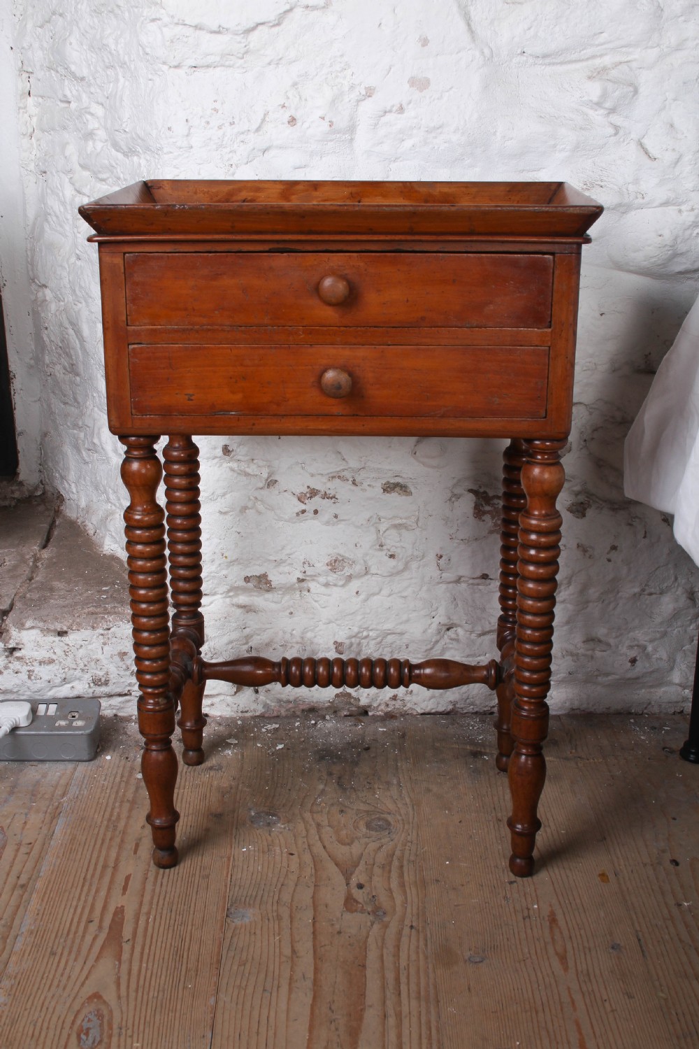 An antique warm brown wooden side table featuring bobbin-turned legs, a tray top, two drawers with round wooden knobs, and a turned stretcher.