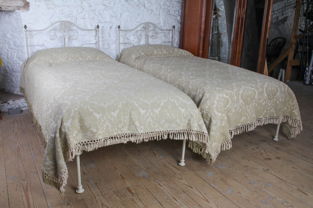 A pair of cream-painted antique Portuguese iron single beds featuring decorative scrolled headboards and low-profile foot ends.