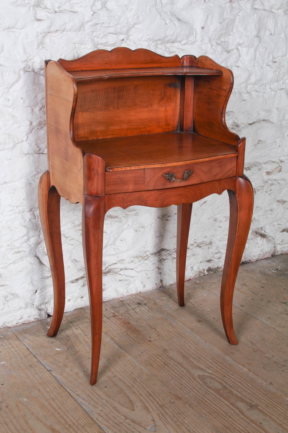 A French Louis XV style cherrywood bedside table featuring slender cabriole legs, a single drawer with a brass handle, and an upper gallery shelf.