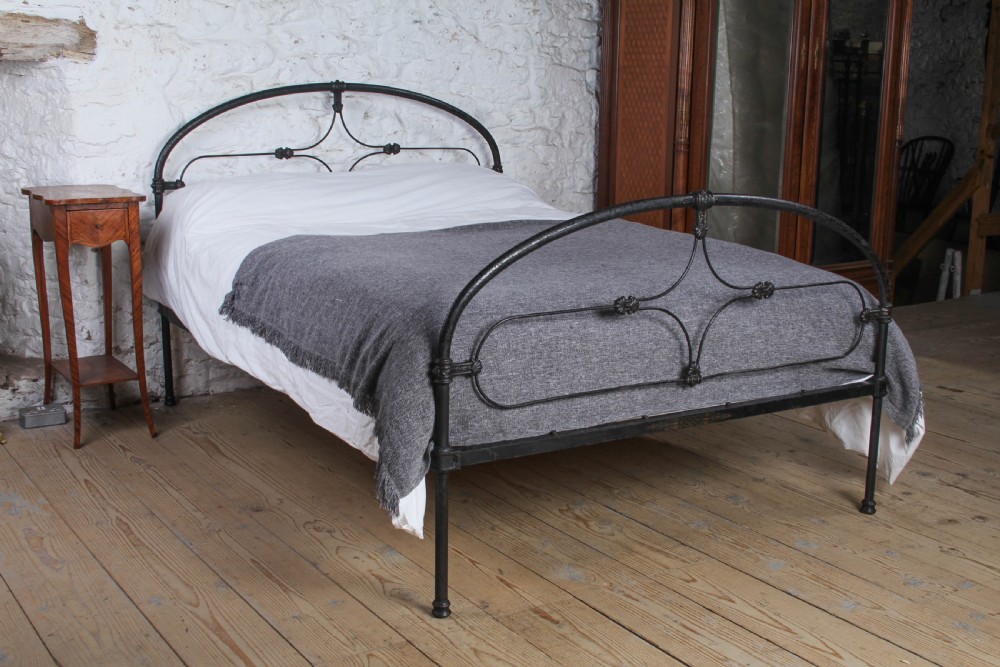 An antique dark wrought iron double bed frame featuring arched headboard and footboard rails with decorative cast rosettes.