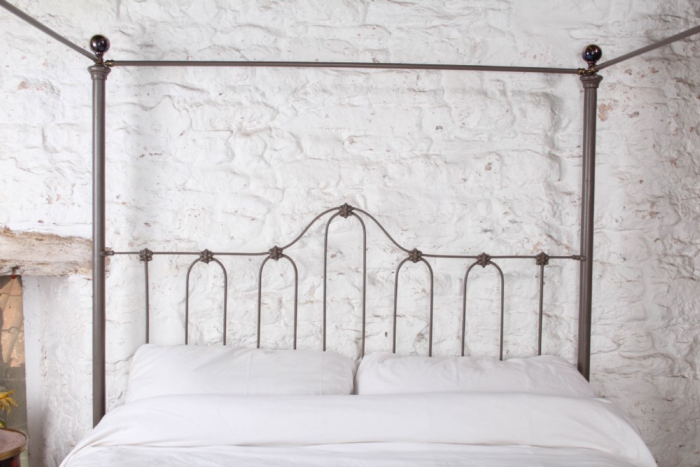 A low height king size metal four poster bed frame with slender corner posts, brass finials, and an arched headboard with vertical iron spindles. View IV