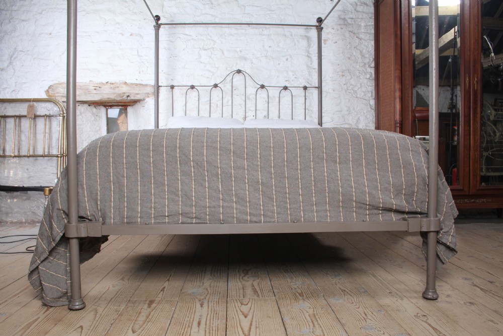 A low height king size metal four poster bed frame with slender corner posts, brass finials, and an arched headboard with vertical iron spindles. View VI