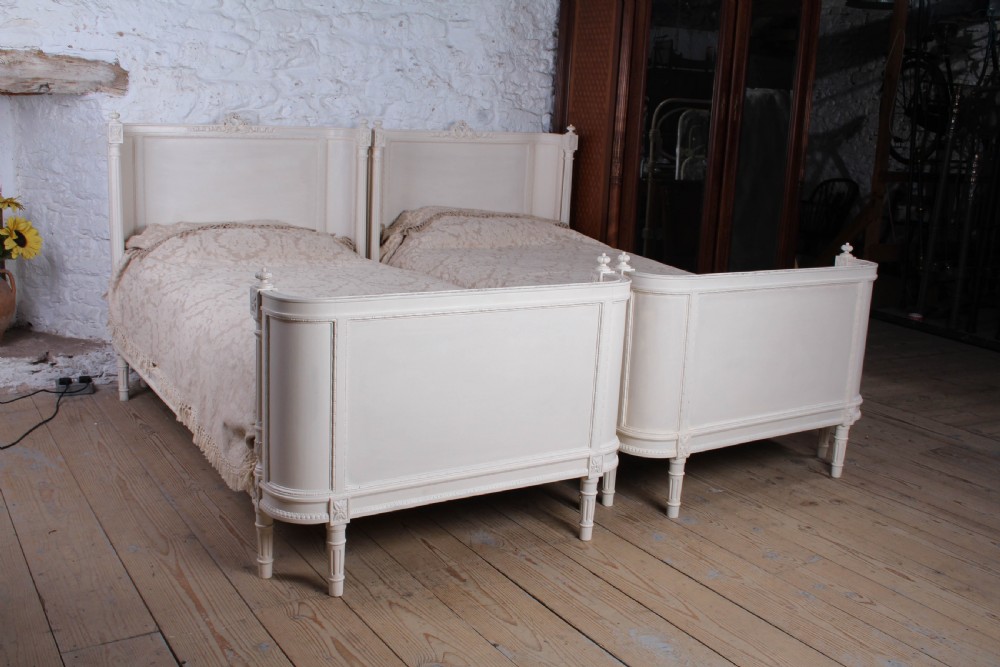 A pair of off-white painted French corbeille single bed frames featuring curved footboards, carved details, and fluted legs.