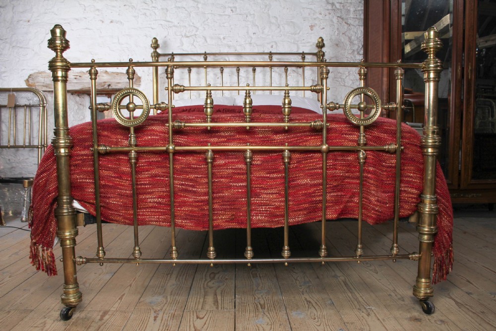 A Victorian brass king-size bed frame featuring turned upright posts, urn finials, vertical decorative spindles, circular roundels, and original castors. View IV