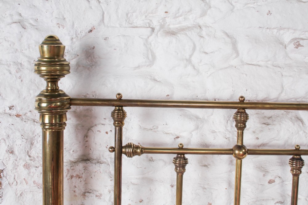 A Victorian brass king-size bed frame featuring turned upright posts, urn finials, vertical decorative spindles, circular roundels, and original castors. View VII