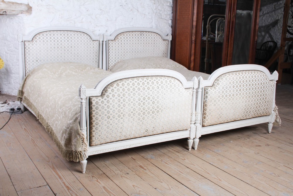 A pair of off-white painted French Empire style single bed frames featuring upholstered arched headboards and matching footboards with geometric fabric.