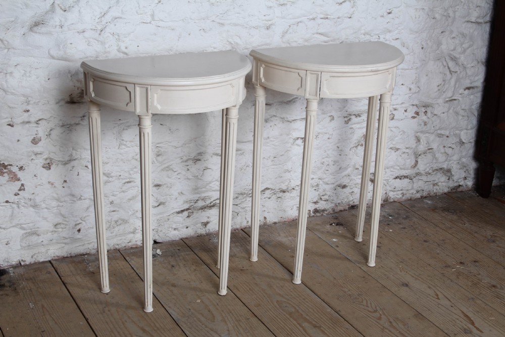 A pair of off-white painted demi-lune console tables with panelled aprons and slender fluted turned legs.
