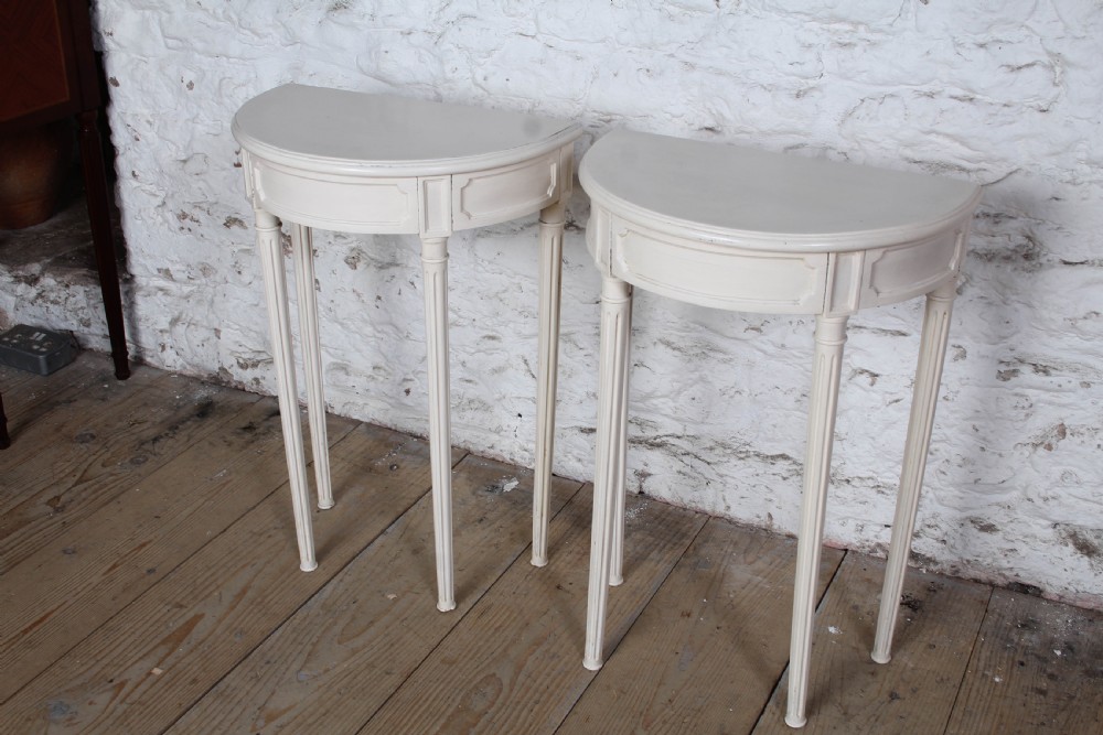 A pair of off-white painted demi-lune console tables with panelled aprons and slender fluted turned legs. View II