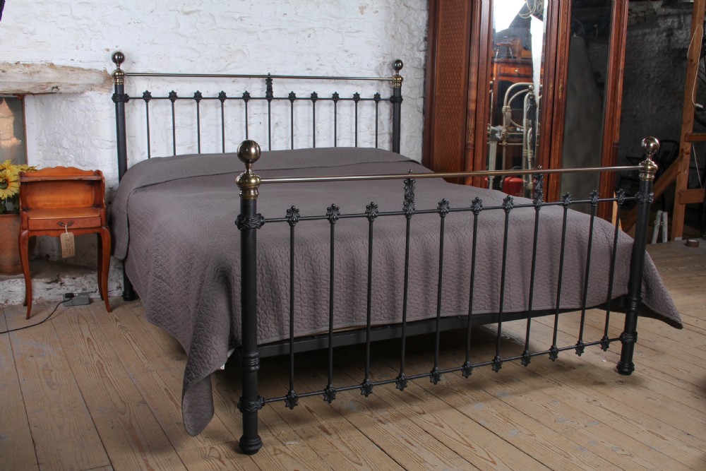 A super king size bedstead featuring a black iron frame with vertical spindles, cast rosettes, and brass top rails with brass ball finials.