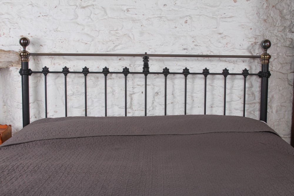 A super king size bedstead featuring a black iron frame with vertical spindles, cast rosettes, and brass top rails with brass ball finials. View VI