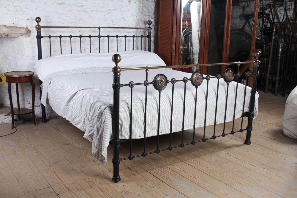 A Victorian black iron and brass super king-size bedstead featuring vertical spindles, circular brass medallions on the footboard, and brass top rails with spherical finials.