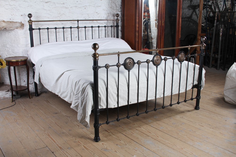 A Victorian black iron and brass super king-size bedstead featuring vertical spindles, circular brass medallions on the footboard, and brass top rails with spherical finials. View II