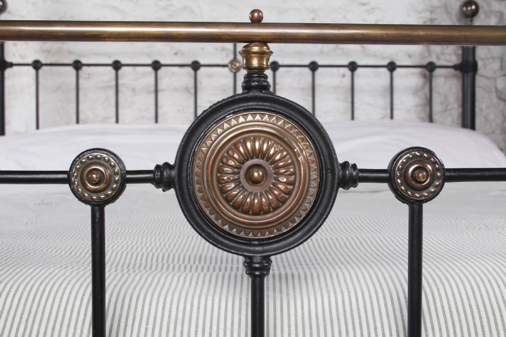 A Victorian black iron and brass super king-size bedstead featuring vertical spindles, circular brass medallions on the footboard, and brass top rails with spherical finials. View V