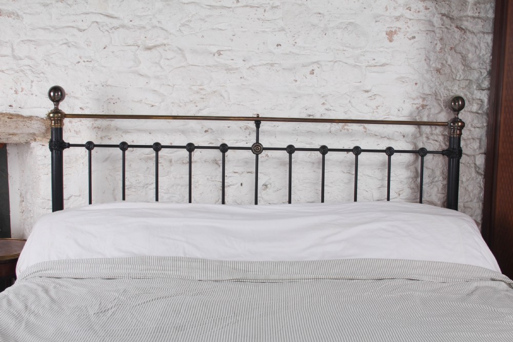 A Victorian black iron and brass super king-size bedstead featuring vertical spindles, circular brass medallions on the footboard, and brass top rails with spherical finials. View IX