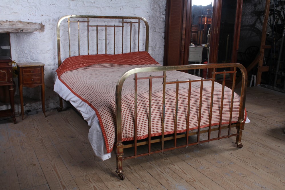 An antique French brass king-size bedstead featuring a square tube frame, rounded top corners, vertical spindles, and brass castors.