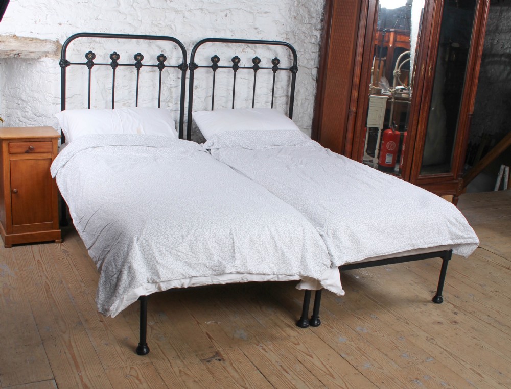 A pair of black wrought iron French single bed frames featuring vertical spindles with cast decorative rosettes and no footboards.