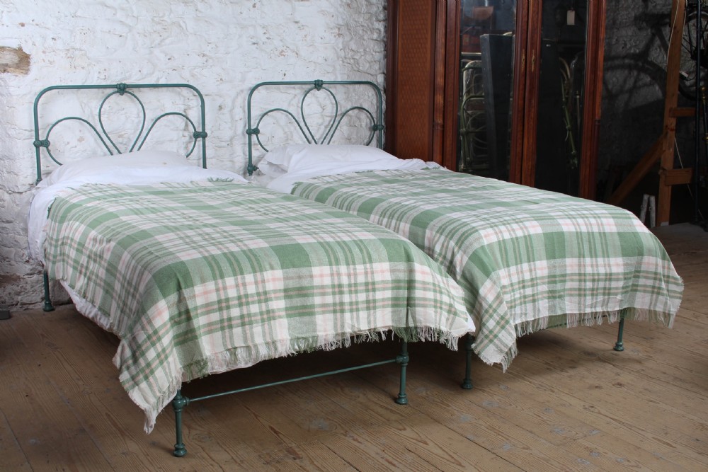 A pair of green painted iron single bed frames featuring scrolled Celtic heart motifs on the headboards and low open ends.