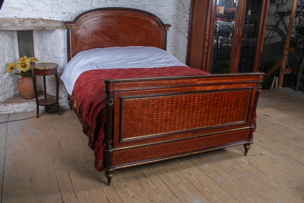 A French king-size bed frame featuring mahogany and fruitwood geometric parquetry inlay, arched headboard, brass trim, and fluted corner posts.