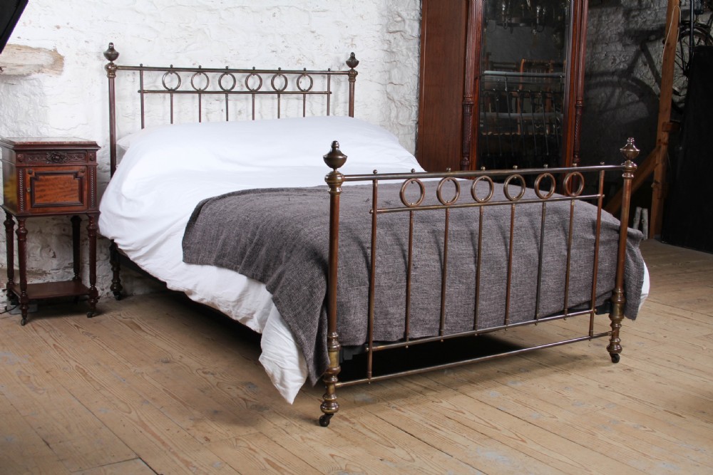A king-size French brass bedstead featuring vertical metal spindles topped with circular ring motifs and decorative urn finials on four corner posts.