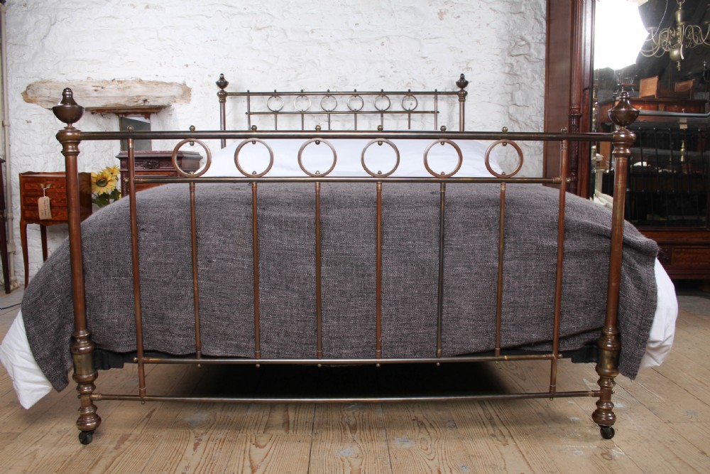 A king-size French brass bedstead featuring vertical metal spindles topped with circular ring motifs and decorative urn finials on four corner posts. View II