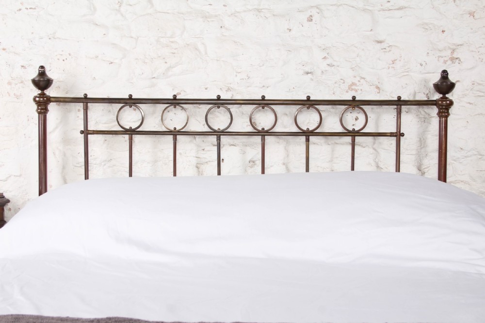 A king-size French brass bedstead featuring vertical metal spindles topped with circular ring motifs and decorative urn finials on four corner posts. View III