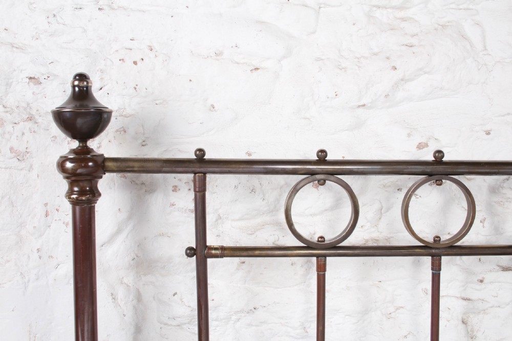 A king-size French brass bedstead featuring vertical metal spindles topped with circular ring motifs and decorative urn finials on four corner posts. View VI