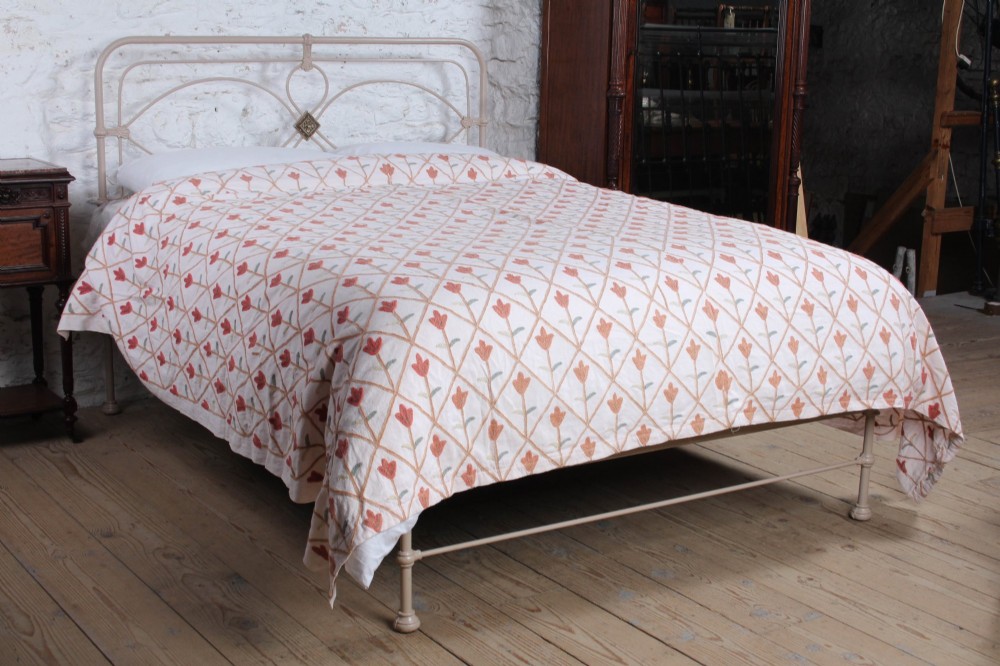 A cream painted wrought iron king size bed frame featuring a high arched headboard with central brass detailing and a low foot end.