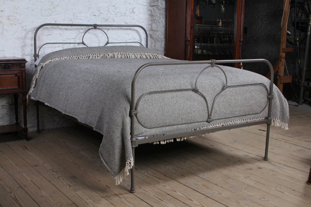 An antique dark grey iron bed frame featuring decorative curved tubing and cast detailing dressed with a textured grey blanket.