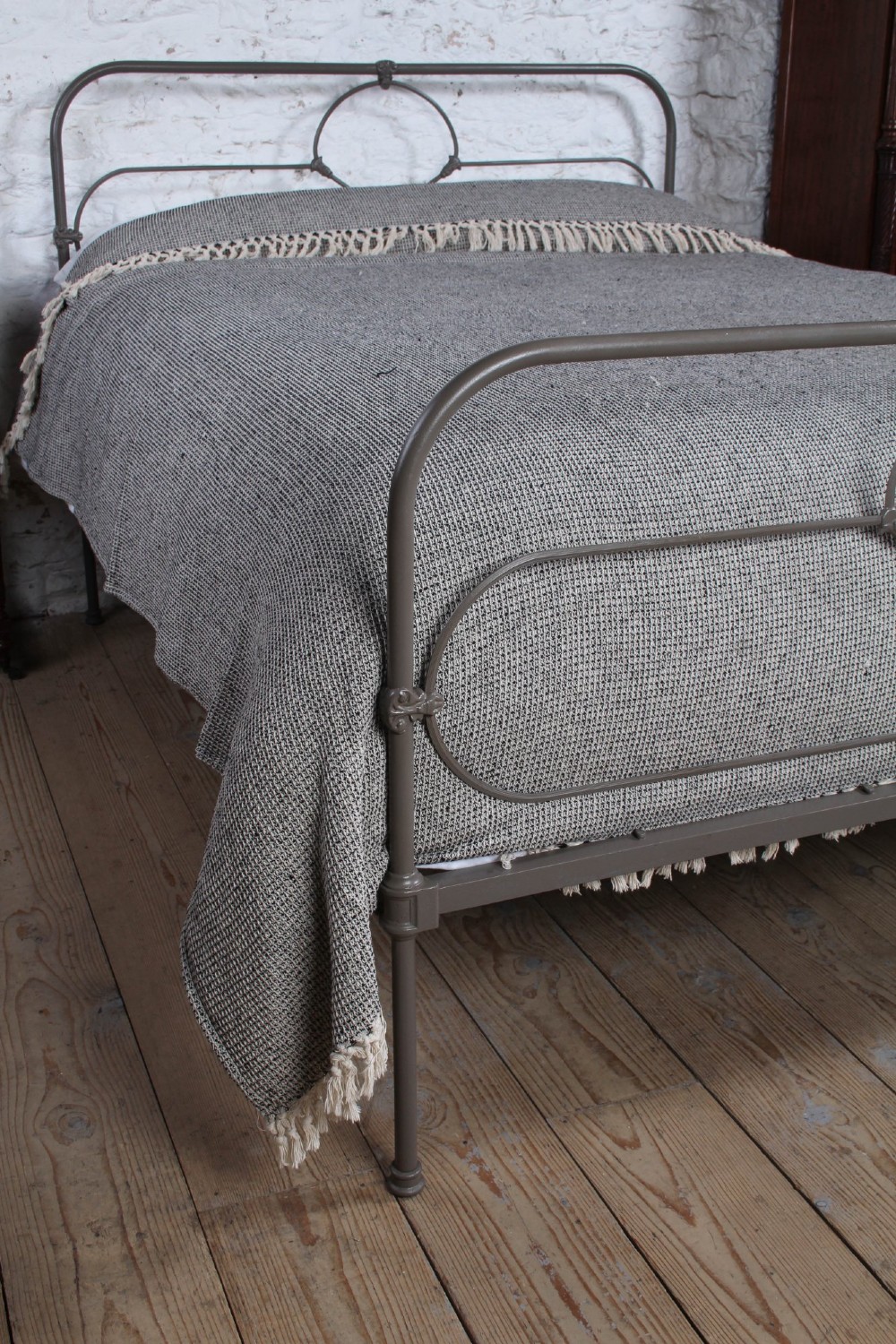 An antique dark grey iron bed frame featuring decorative curved tubing and cast detailing dressed with a textured grey blanket. View III