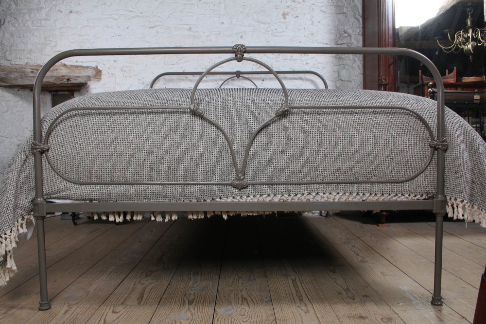 An antique dark grey iron bed frame featuring decorative curved tubing and cast detailing dressed with a textured grey blanket. View V