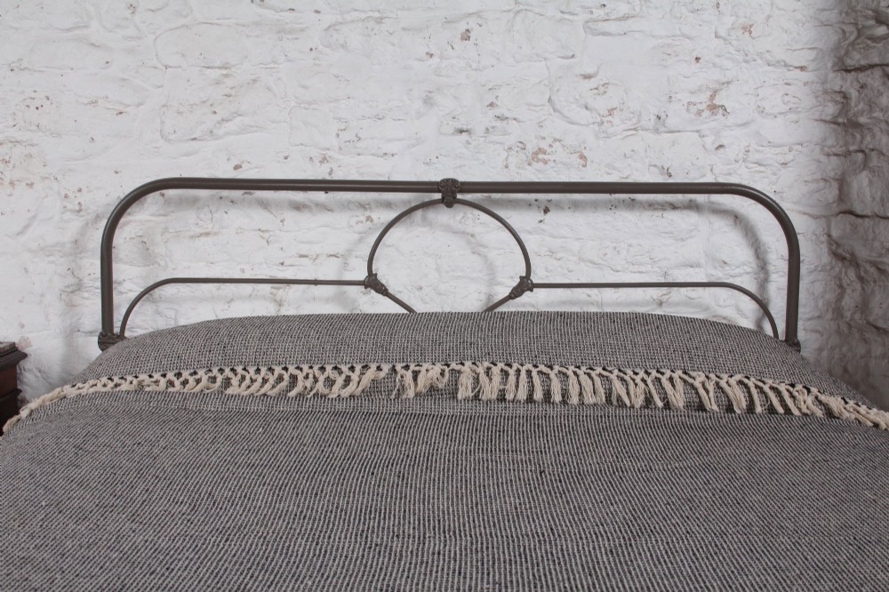 An antique dark grey iron bed frame featuring decorative curved tubing and cast detailing dressed with a textured grey blanket. View VI