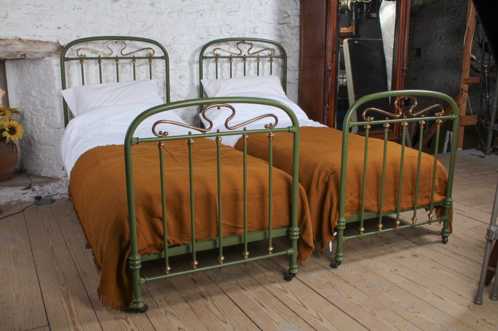 A pair of olive green painted iron single bed frames featuring brass finials, vertical spindles, and decorative scrolled crests.