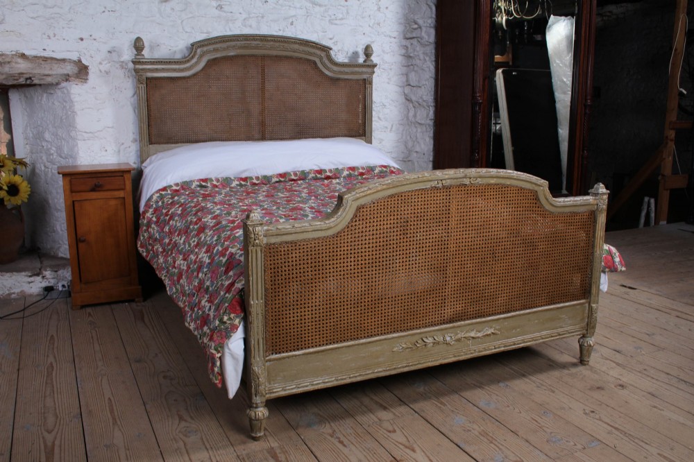 A French Louis XVI style double bed frame with a carved wood surround, woven cane headboard and footboard, fluted legs, and a distressed grey painted finish.