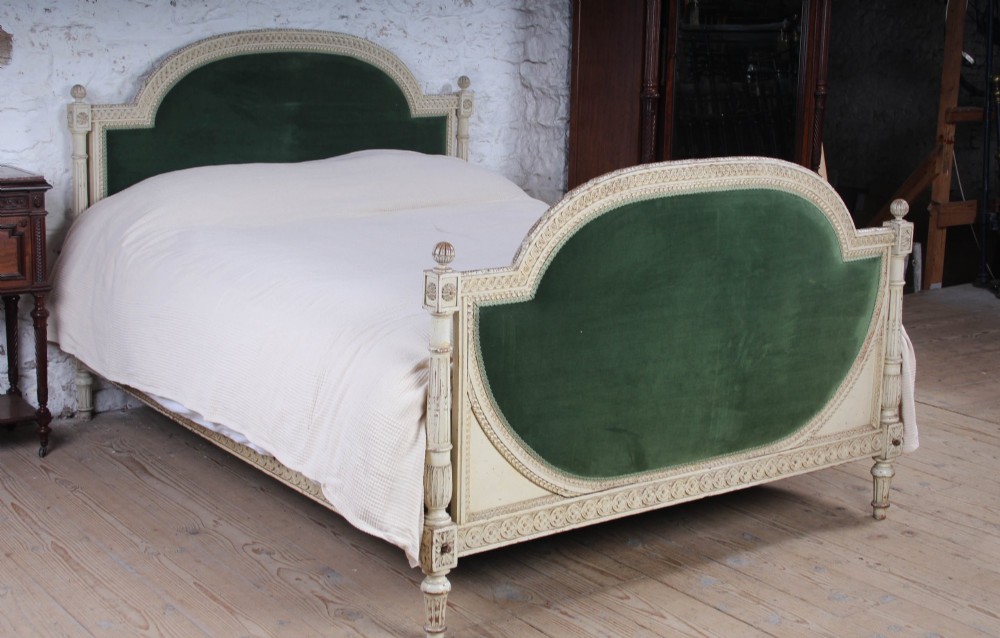 An antique French Louis XVI style bedstead featuring a cream-painted carved wooden frame, fluted posts, finials, and arched headboard and footboard panels upholstered in green velvet.