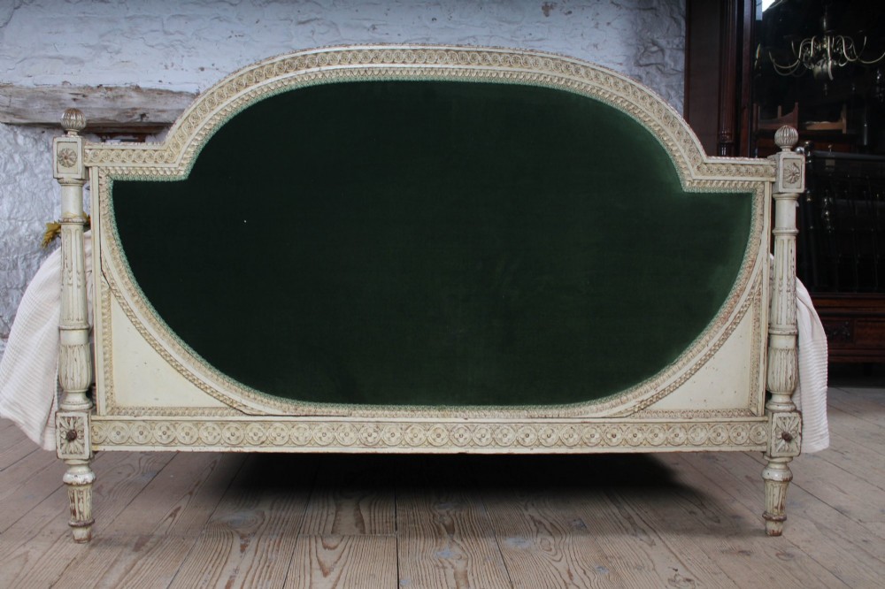 An antique French Louis XVI style bedstead featuring a cream-painted carved wooden frame, fluted posts, finials, and arched headboard and footboard panels upholstered in green velvet. View II