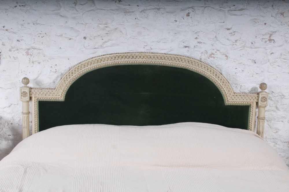 An antique French Louis XVI style bedstead featuring a cream-painted carved wooden frame, fluted posts, finials, and arched headboard and footboard panels upholstered in green velvet. View VI