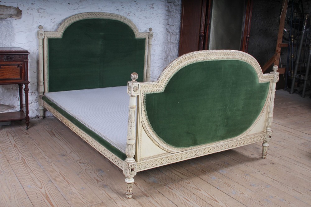 An antique French Louis XVI style bedstead featuring a cream-painted carved wooden frame, fluted posts, finials, and arched headboard and footboard panels upholstered in green velvet. View VIII