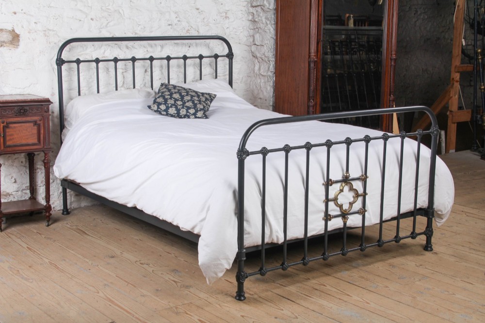 A black iron and brass king-size bed frame featuring vertical spindles and a central brass quatrefoil motif on the footboard.