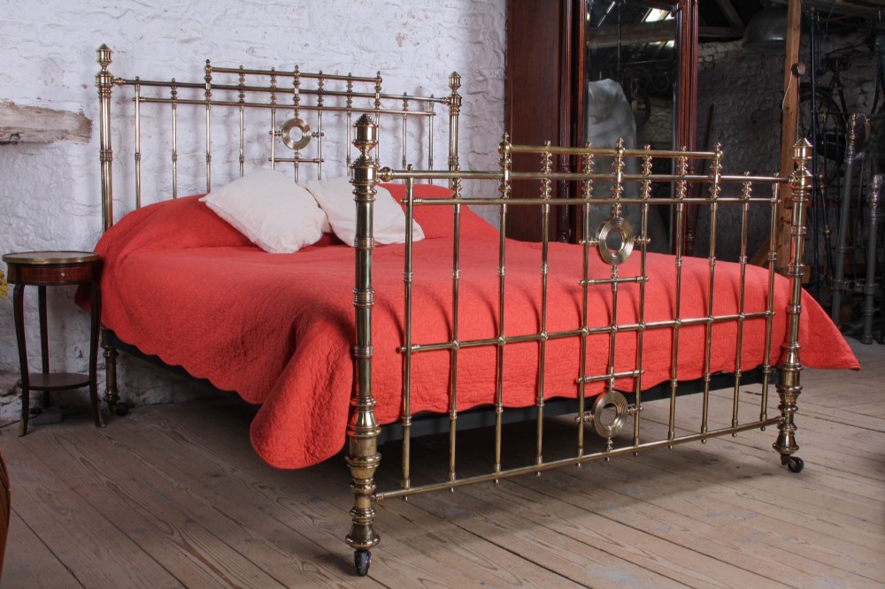 An ornate Victorian polished brass super king bedstead featuring turned posts, decorative circular motifs, vertical spindles, and castors.
