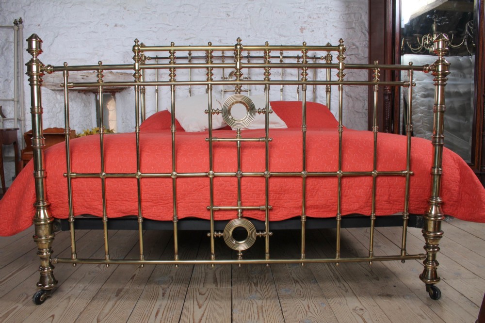 An ornate Victorian polished brass super king bedstead featuring turned posts, decorative circular motifs, vertical spindles, and castors. View II