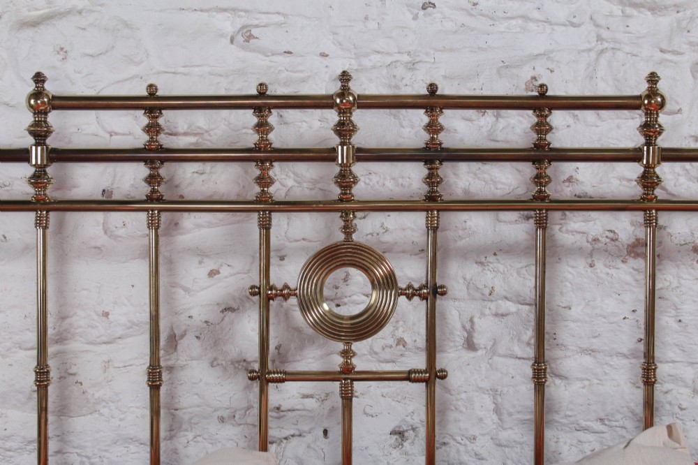 An ornate Victorian polished brass super king bedstead featuring turned posts, decorative circular motifs, vertical spindles, and castors. View X