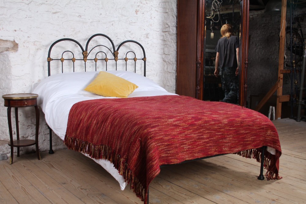 A black Victorian cast iron double bedstead featuring triple arched headboard metalwork with brass castings and a low foot end.